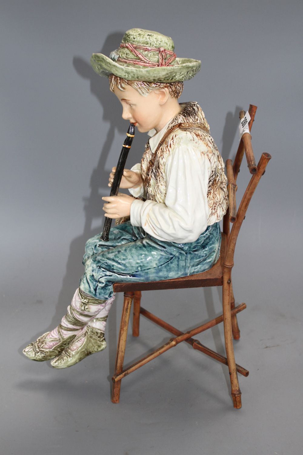 A late 19th century Austrian tin glaze pottery figure of a Tyrolean boy playing a flute, seated upon a bamboo wood chair, height 43cm